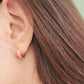 Close-up of a woman’s ear wearing pink and gold enamel huggie earrings, showcasing their smooth finish and playful wavy design.