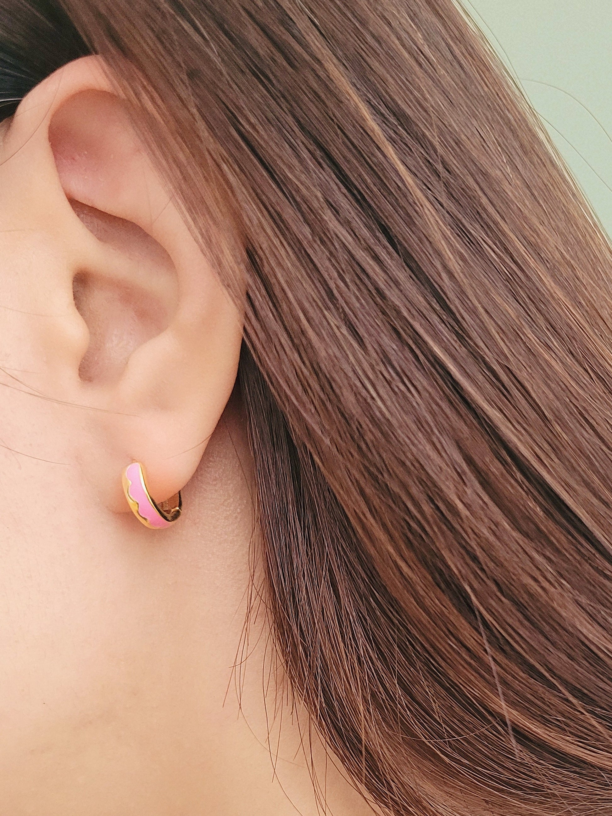 Close-up of a woman’s ear wearing pink and gold enamel huggie earrings, showcasing their smooth finish and playful wavy design.