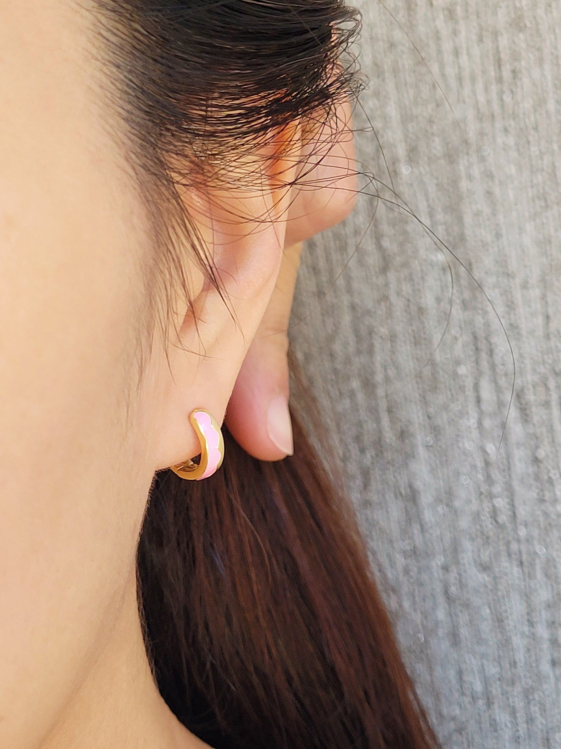 Close-up of a woman’s ear wearing pink and gold enamel huggie earrings, showcasing their smooth finish and playful wavy design.