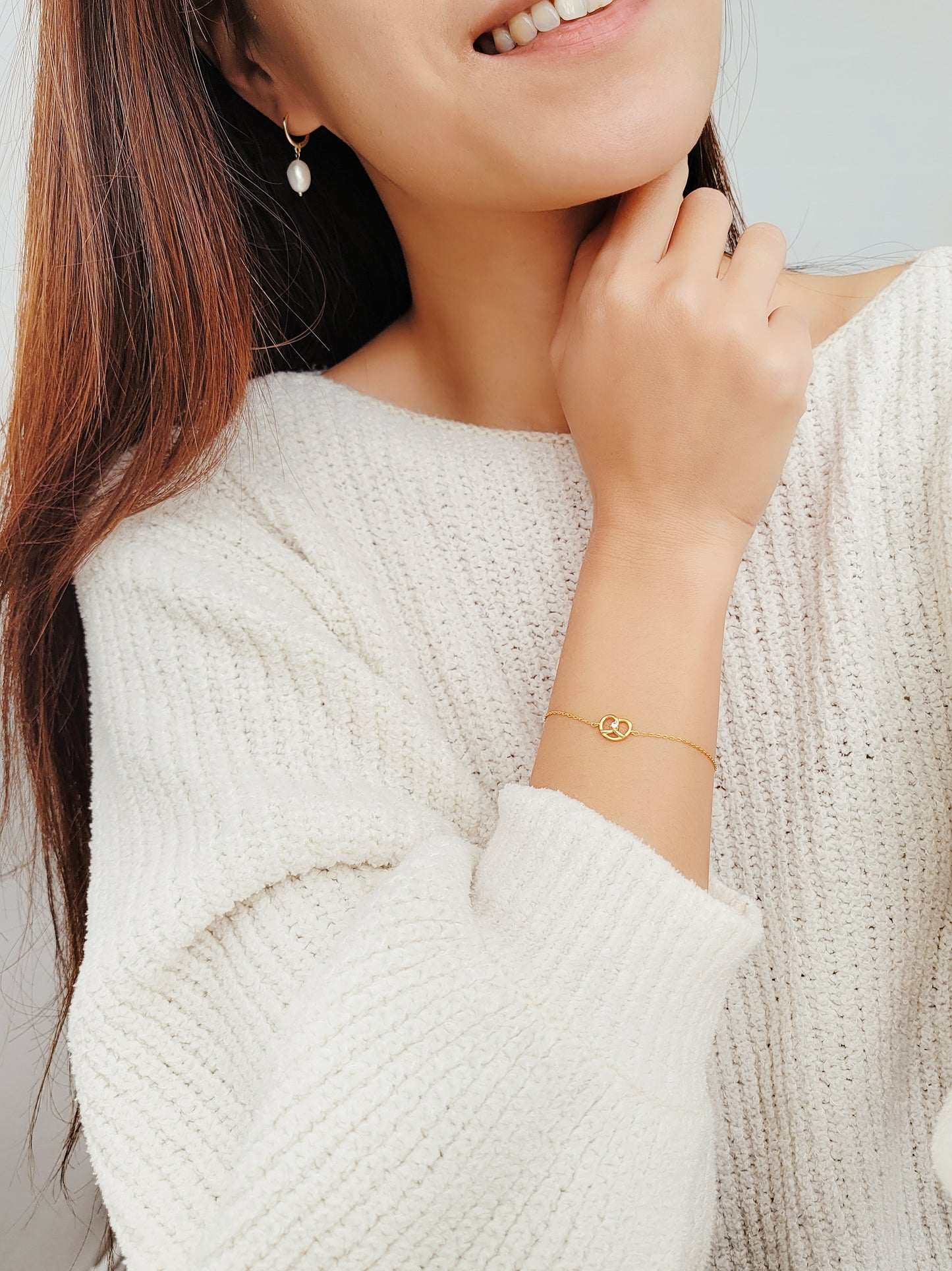 A woman wearing a white sweater, proudly displaying her charming gold pretzel bracelet.