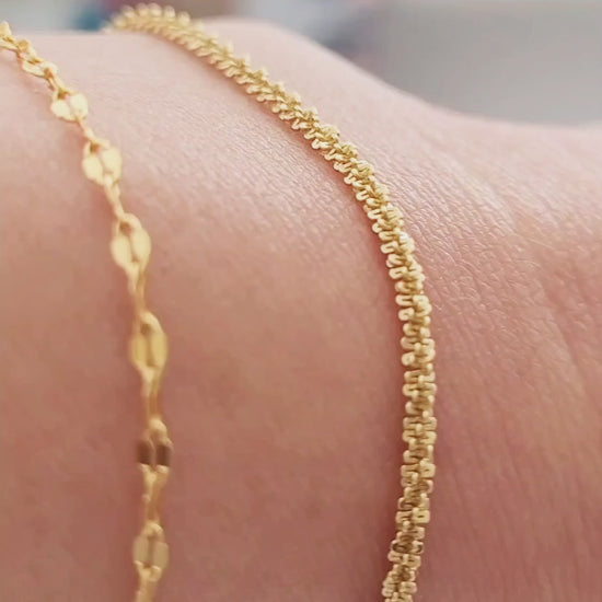 Close-up of a woman resting her arm on her body, wearing a delicate, shimmering gold rope bracelet against her wrist, with a cozy, knit sweater in the background.