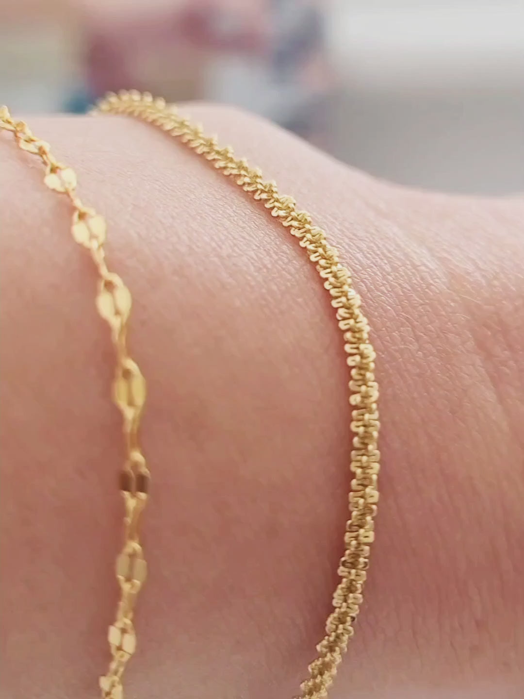 Close-up of a woman resting her arm on her body, wearing a delicate, shimmering gold rope bracelet against her wrist, with a cozy, knit sweater in the background.