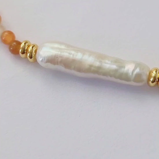 Close-up of a woman wearing an orange agate beaded necklace with a central baroque pearl. The necklace features small, round orange beads accented by gold elements and a single elongated, irregular-shaped white pearl in the center. 