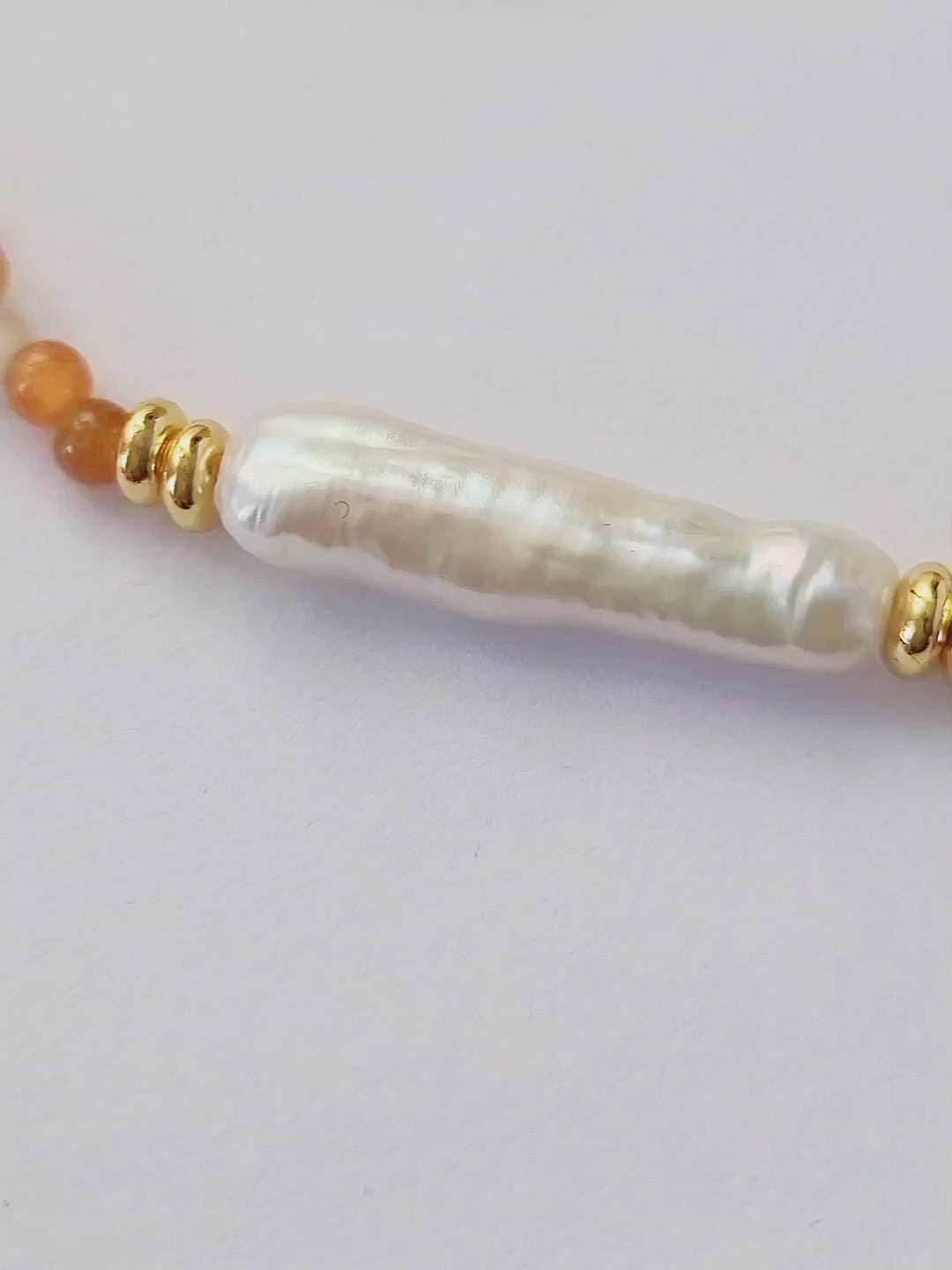 Close-up of a woman wearing an orange agate beaded necklace with a central baroque pearl. The necklace features small, round orange beads accented by gold elements and a single elongated, irregular-shaped white pearl in the center. 