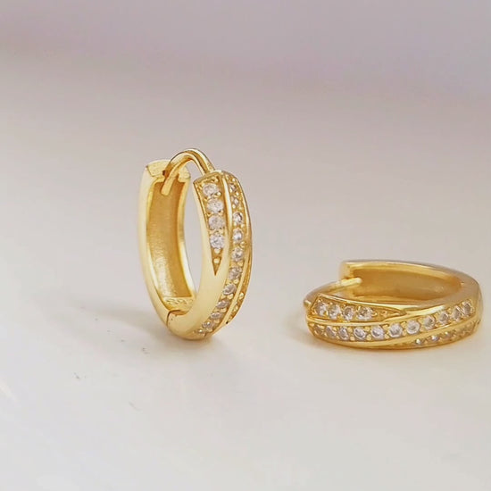 Close-up of two gold crisscross huggie hoop earrings being held between fingers. The earrings feature a delicate design with intersecting bands, both adorned with small, sparkling stones.