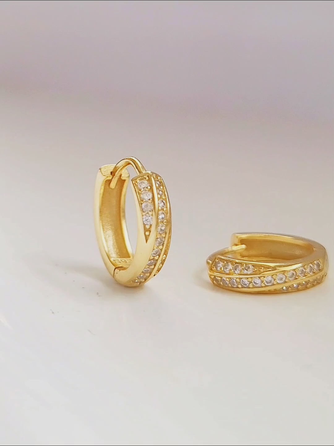 Close-up of two gold crisscross huggie hoop earrings being held between fingers. The earrings feature a delicate design with intersecting bands, both adorned with small, sparkling stones.