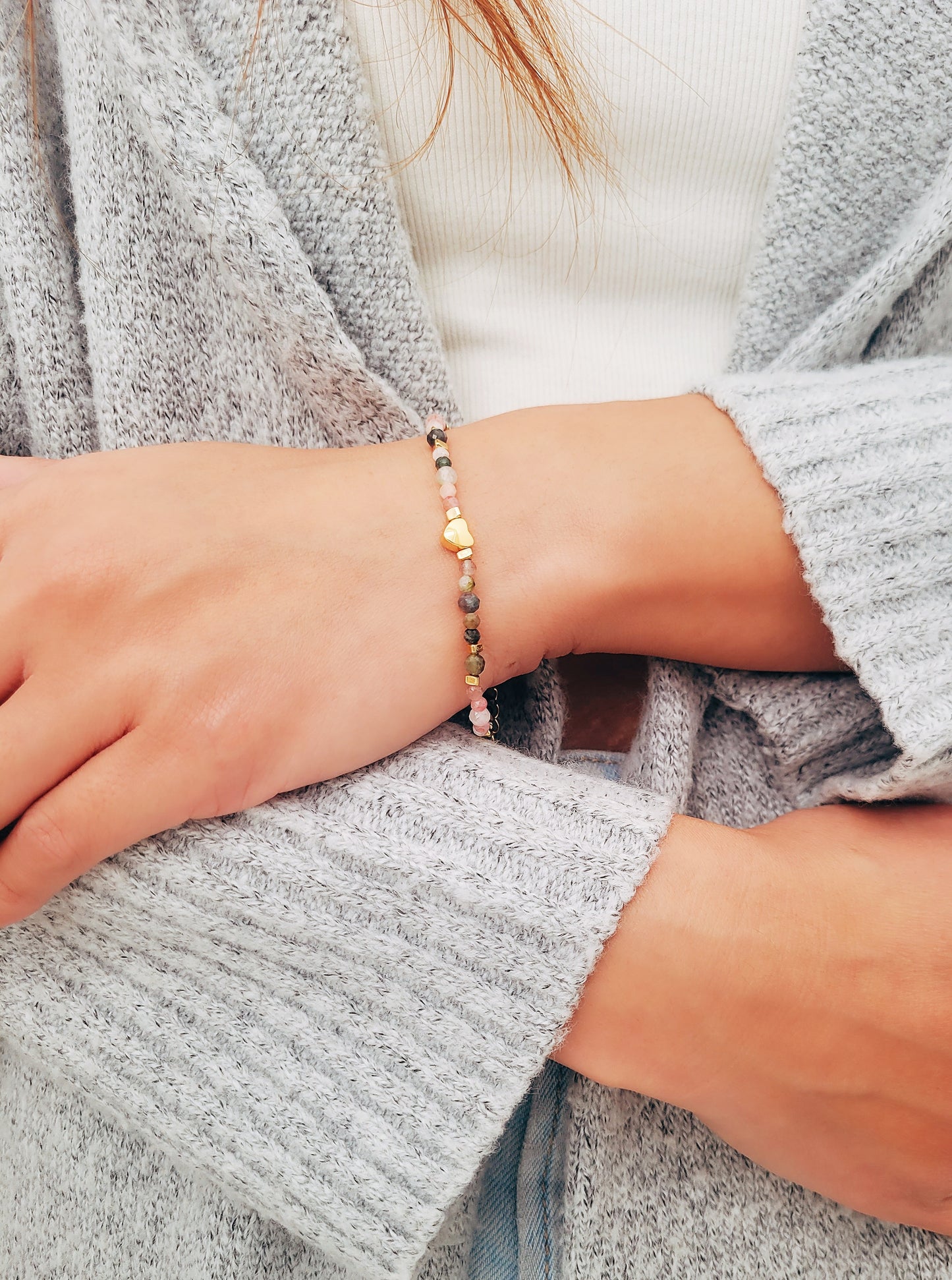 A woman wearing a dainty rainbow gemstone bracelet featuring faceted aventurine, jasper, and rose quartz beads, accented with gold vermeil heart charm at the center. The bracelet has a gold clasp and adjustable chain.