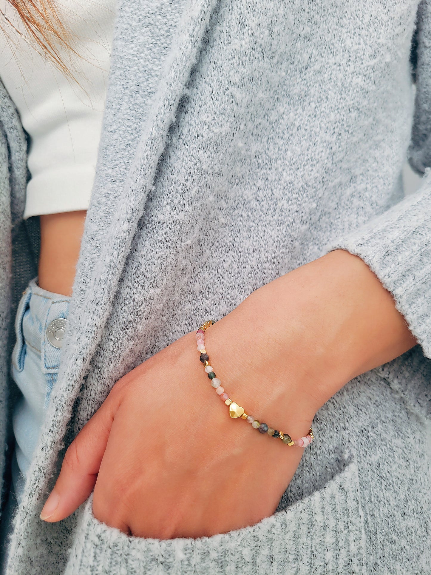 A stylish woman wearing a dainty rainbow gemstone bracelet featuring faceted aventurine, jasper, and rose quartz beads, accented with gold vermeil heart charm at the center. The bracelet has a gold clasp and adjustable chain.