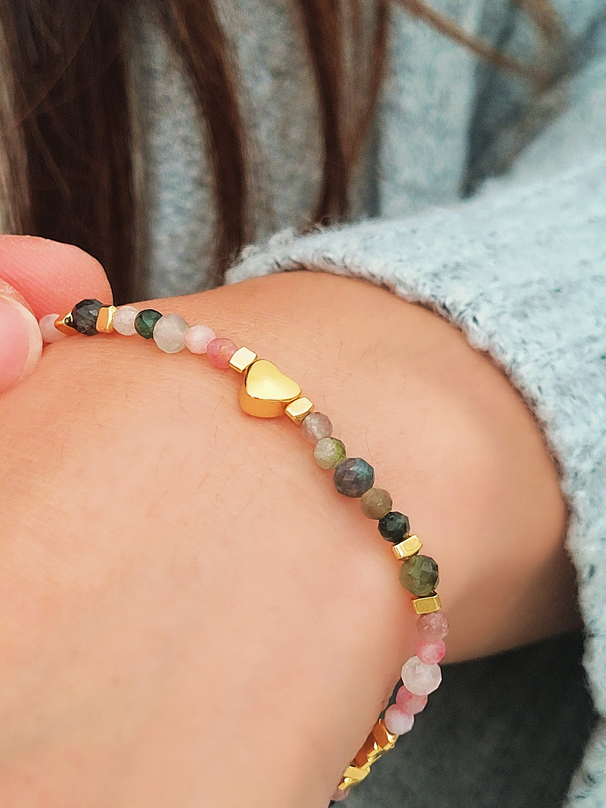 Close-up of a woman's waist wearing a dainty rainbow gemstone bracelet featuring faceted aventurine, jasper, and rose quartz beads, accented with gold vermeil heart charm at the center. The bracelet has a gold clasp and adjustable chain.