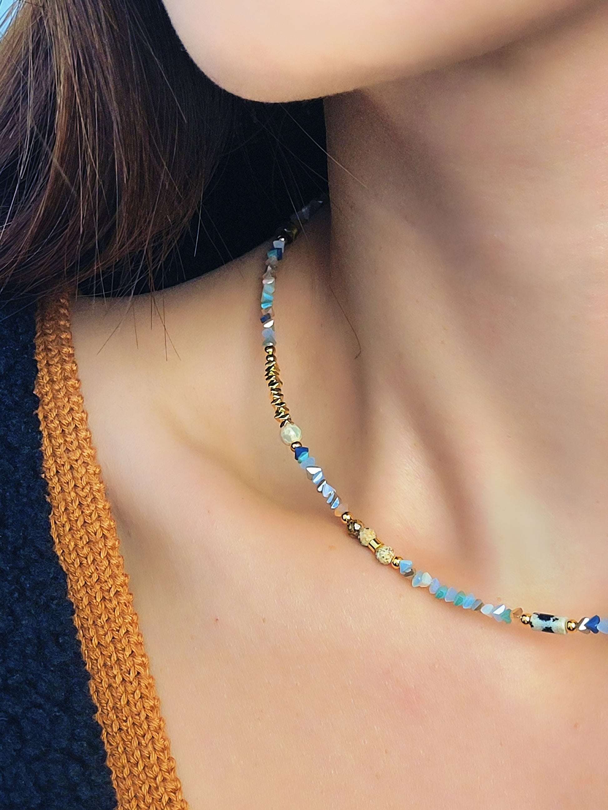 Close-up of a woman wearing a delicate multi-gemstone necklace featuring small, irregular-shaped beads in various shades of  earthy color palette with gold accents and natural stones, creating a minimalistic yet bohemian aesthetic.