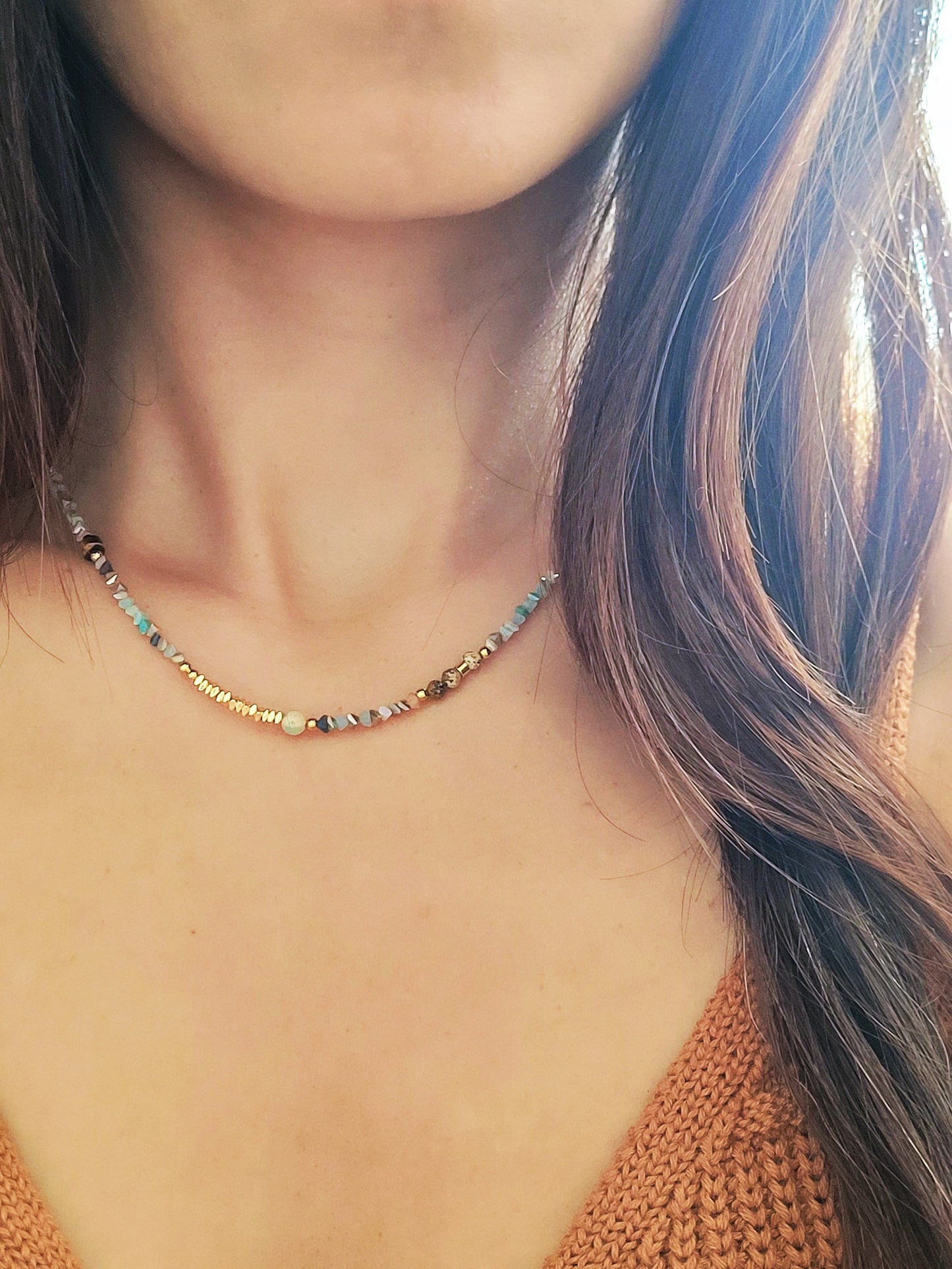 Close-up of a woman wearing a delicate multi-gemstone necklace featuring small, irregular-shaped beads in various shades of blue, gold, and brown. The necklace has a subtle, earthy color palette with gold accents and natural stones, creating a minimalistic yet bohemian aesthetic. The woman is dressed in a light orange knit top, with her hair resting on one side.