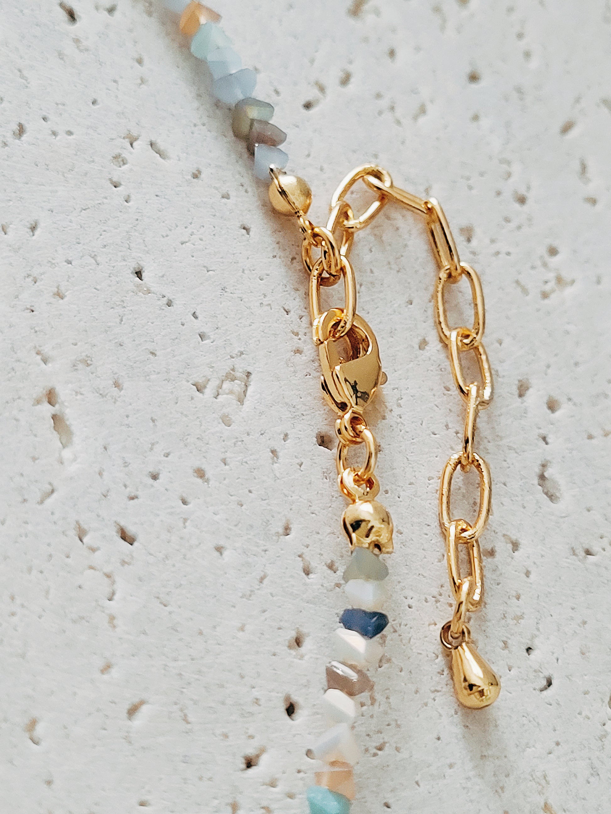 Close-up of a delicate multi-gemstone necklace featuring small, irregular-shaped beads in various shades of blue, gold, and brown. The necklace has a subtle, earthy color palette with gold accents and natural stones, creating a minimalistic yet bohemian aesthetic. 