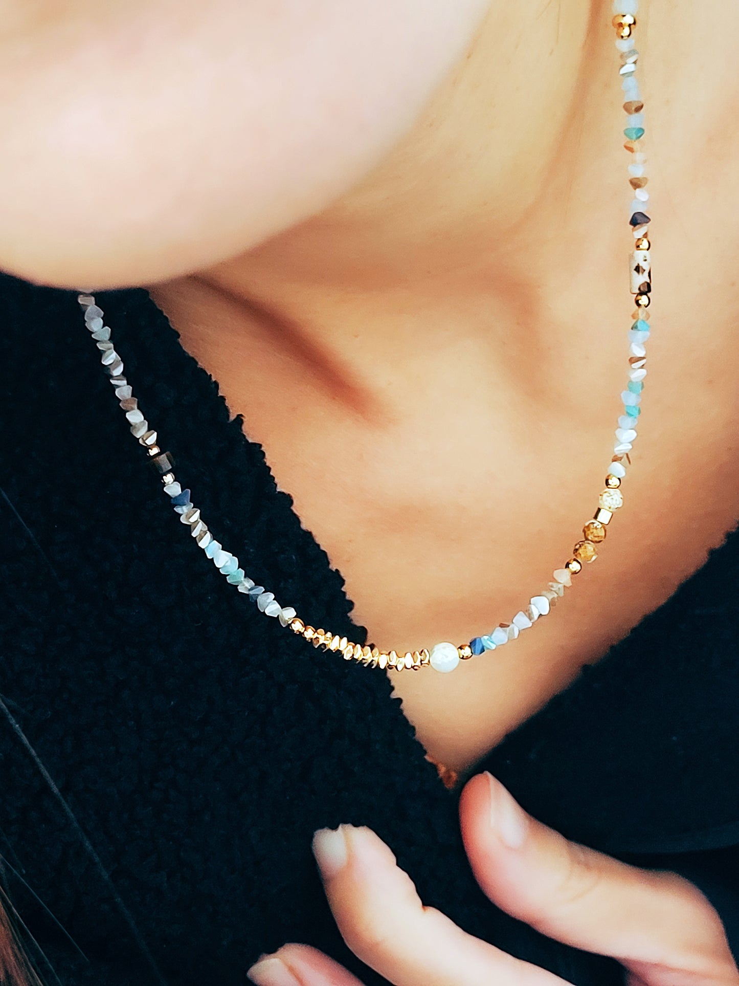 Close-up of a woman wearing a delicate multi-gemstone necklace featuring small, irregular-shaped beads in various shades of blue, gold, and brown. The necklace has a subtle, earthy color palette with gold accents and natural stones, creating a minimalistic yet bohemian aesthetic. The woman is dressed in a light orange knit top, with her hair resting on one side.