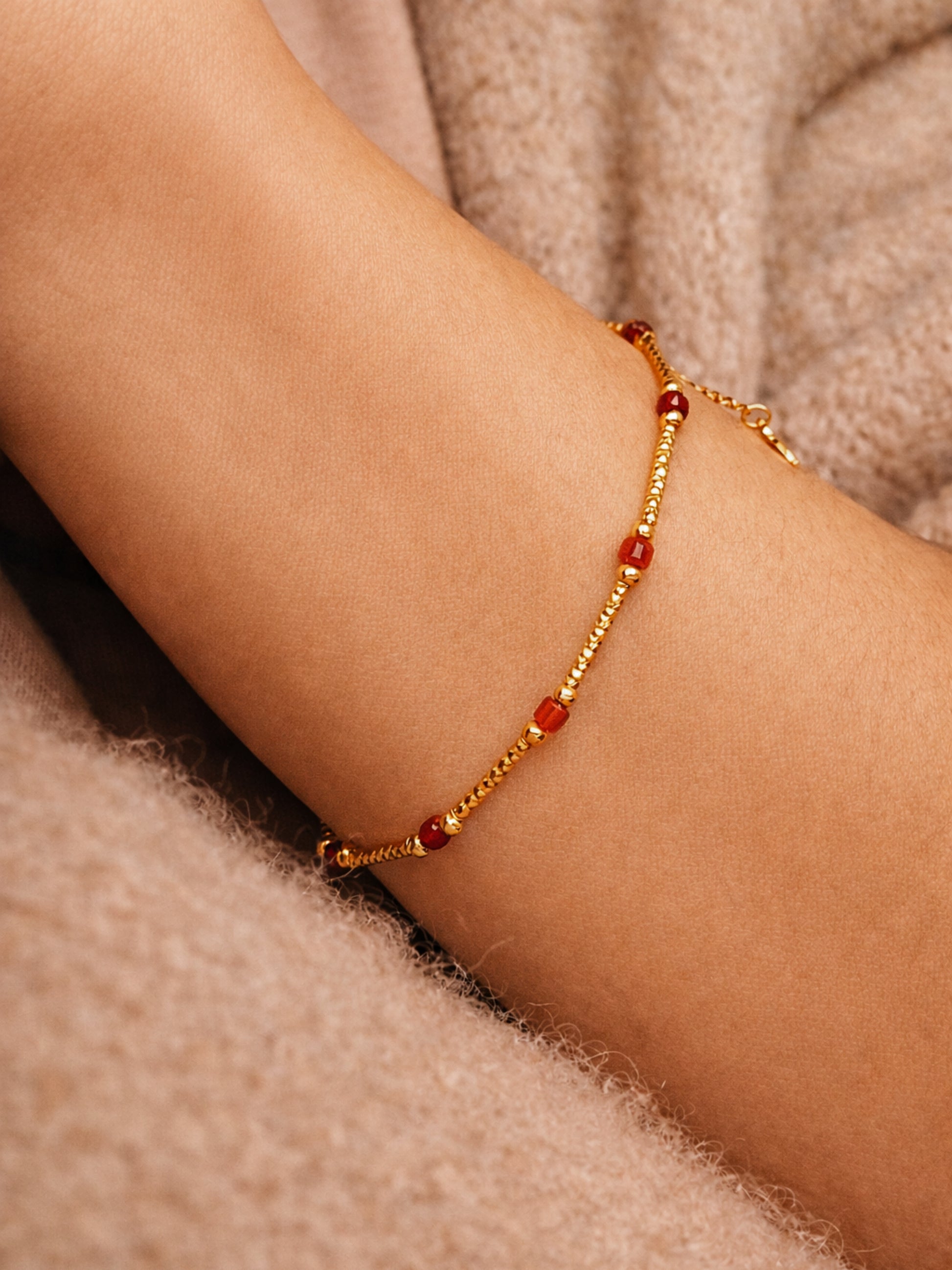 Close-up of a woman's arm wearing a delicate bracelet with red beads on a soft beige background.