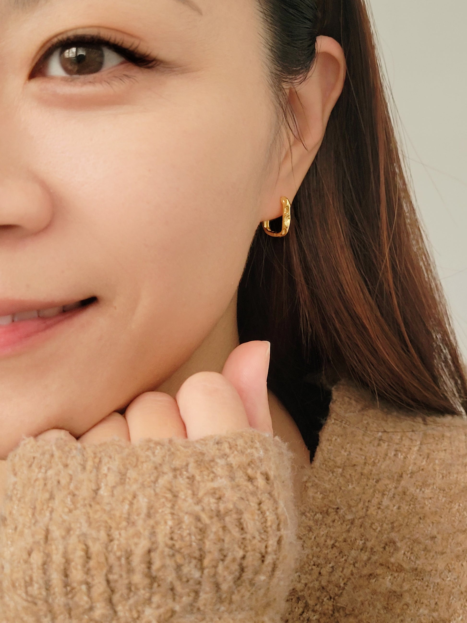 A stylish woman in a brown sweater elegantly wears hammered gold hoop earrings, embodying grace and refined taste.