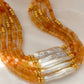 Close-up of a stack of orange agate beaded necklace with a central baroque pearl. The necklace features small, round orange beads accented by gold elements and a single elongated, irregular-shaped white pearl in the center.