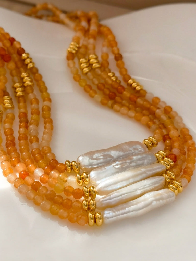 Close-up of a stack of orange agate beaded necklace with a central baroque pearl. The necklace features small, round orange beads accented by gold elements and a single elongated, irregular-shaped white pearl in the center.