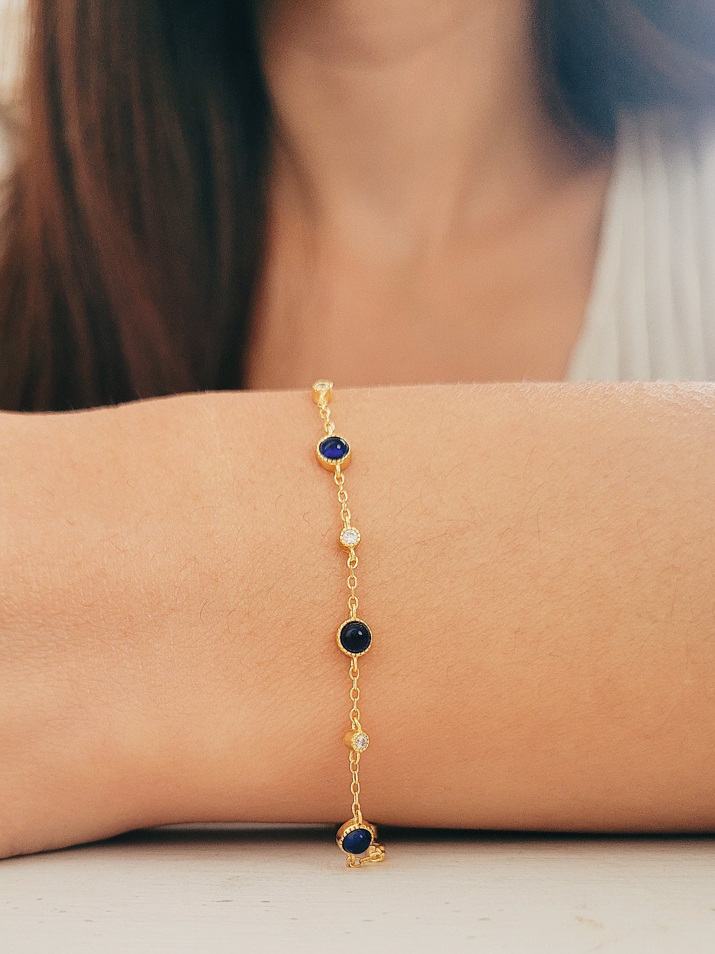 A woman elegantly showcases a sapphire station bracelet adorned with shimmering natural sapphire gemstones and radiant gold plated chain.