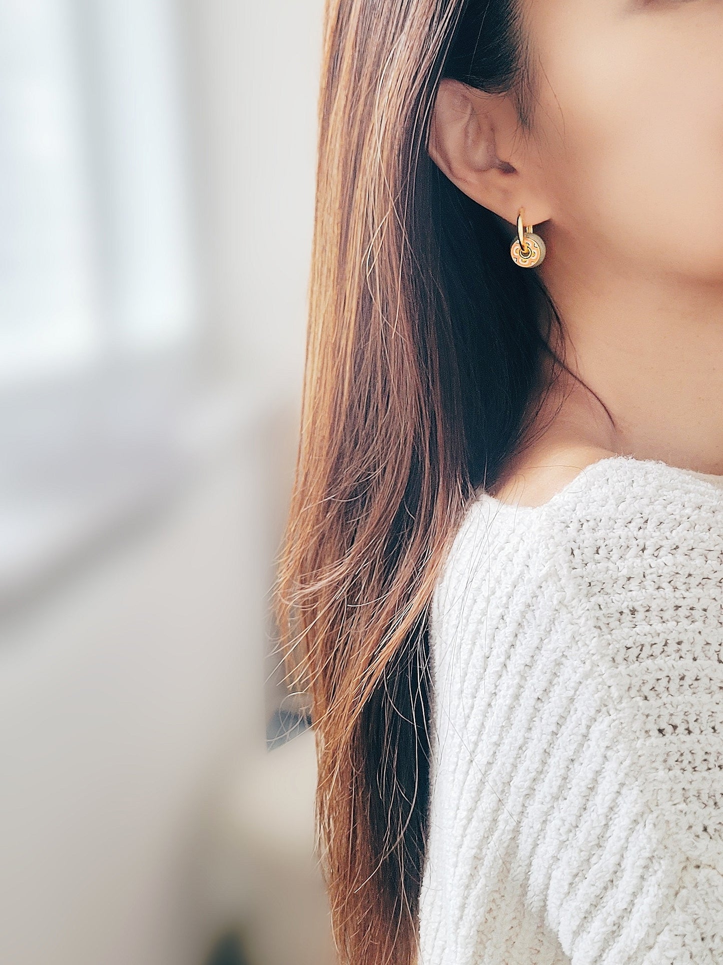 A woman wearing a white sweater and eye-catching green jade drop hoop earrings, set against a vibrant tile pattern.