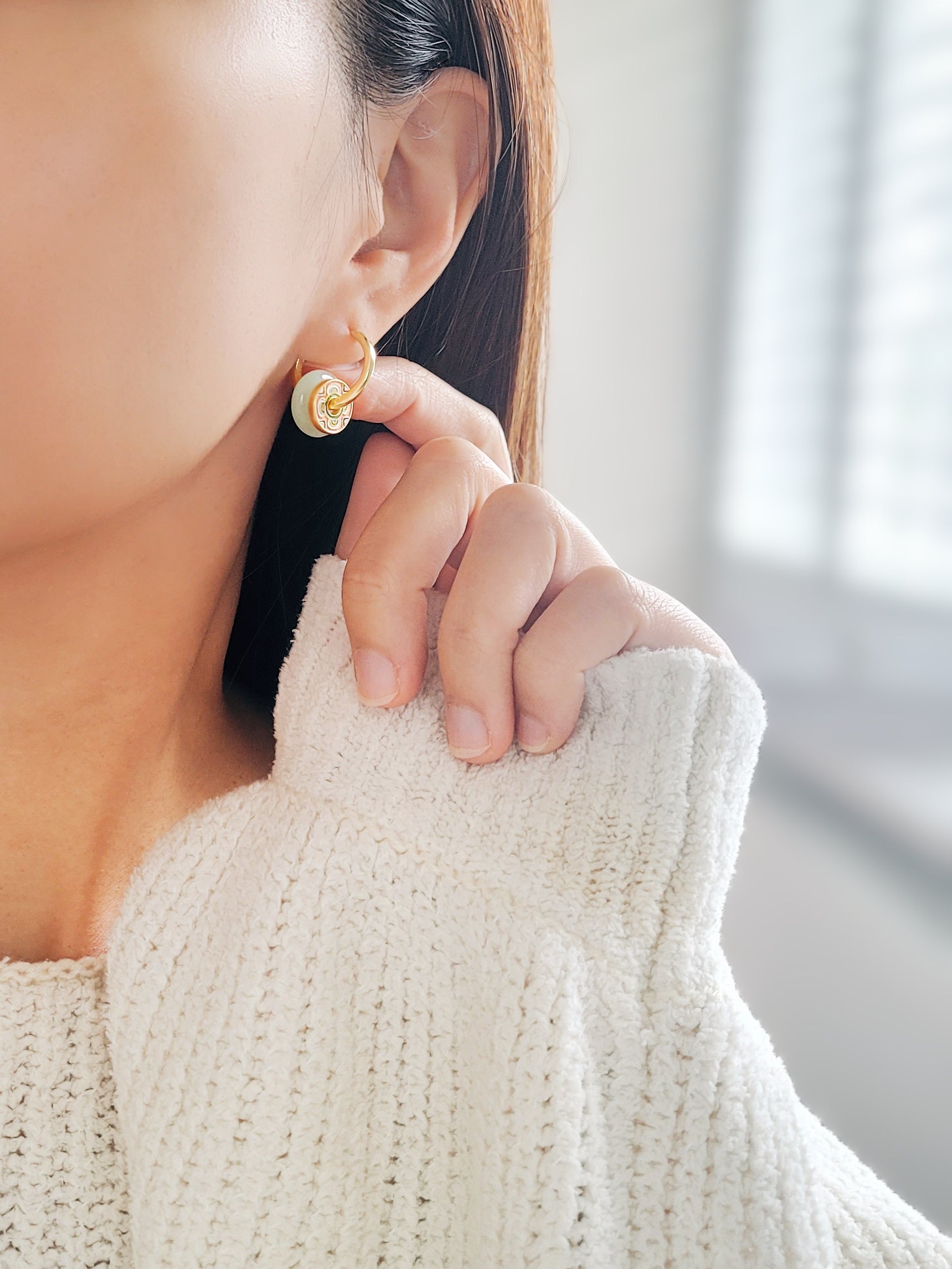 A woman in a cozy white sweater, showcasing stunning green jade drop hoop earrings against a stylish tile pattern.