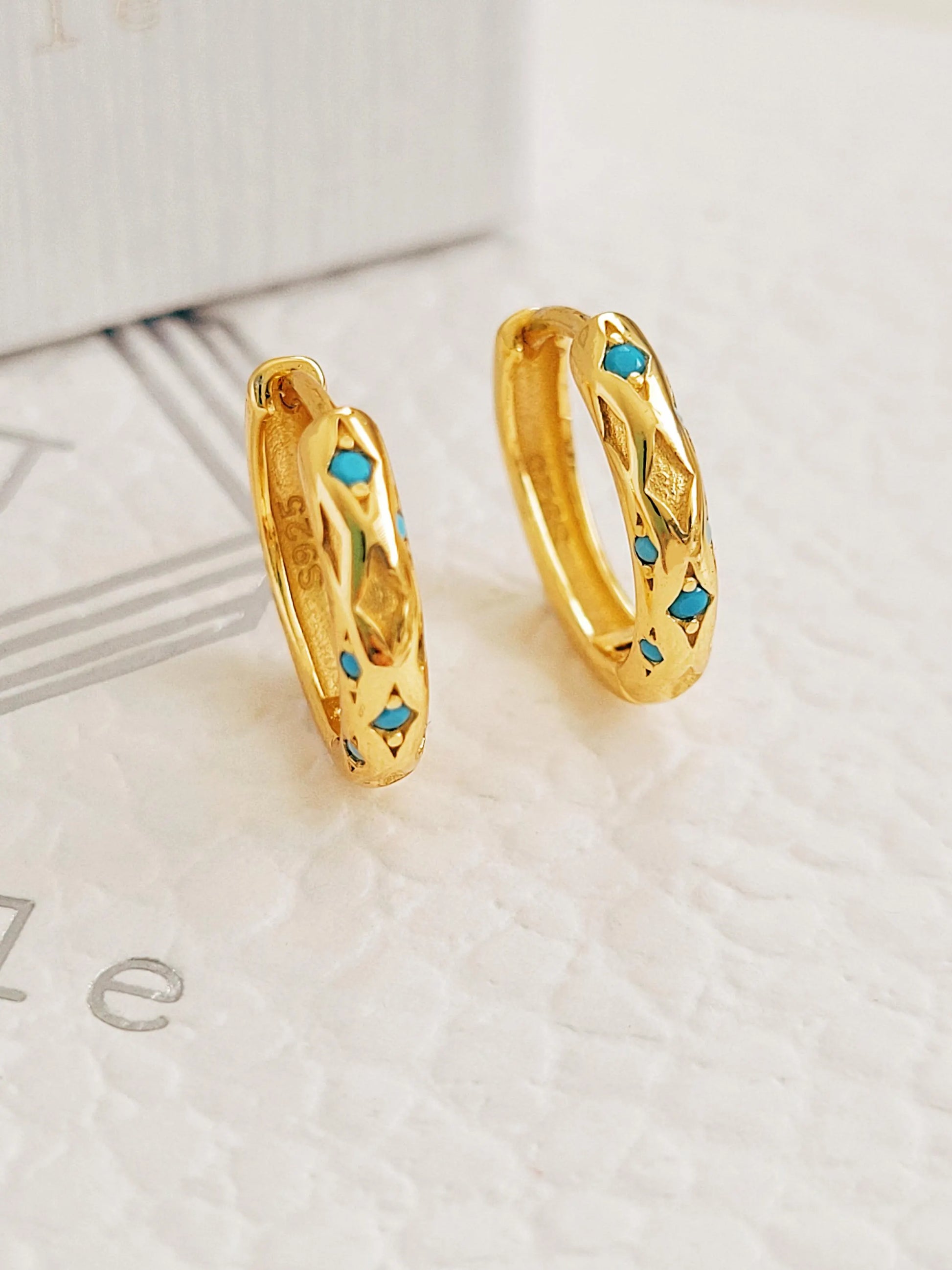 A pair of textured 18K gold-plated over 925 sterling silver huggie earrings featuring engraved star patterns and inlaid vibrant turquoise stones, shown close up.