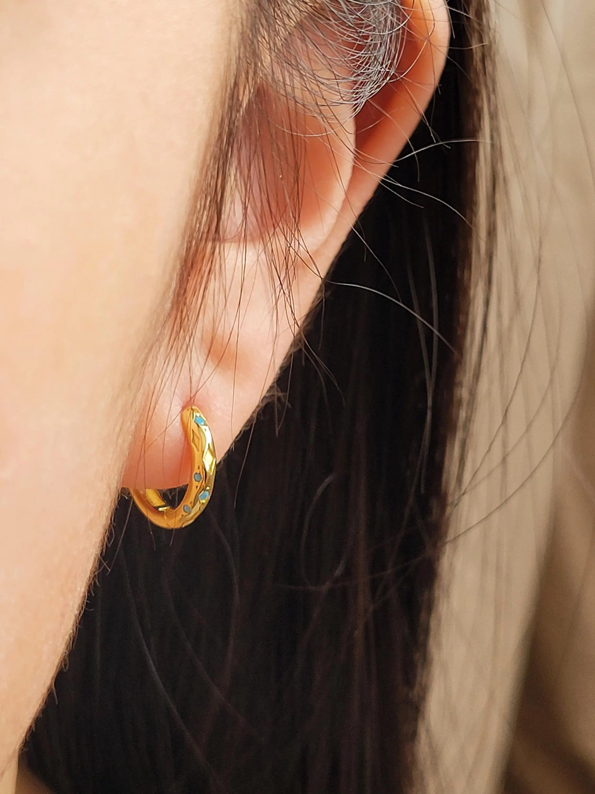 A close-up of a woman's ear displaying a small, dainty 18K gold-plated huggie hoop earring intricately inlaid with tiny turquoise starburst stones.